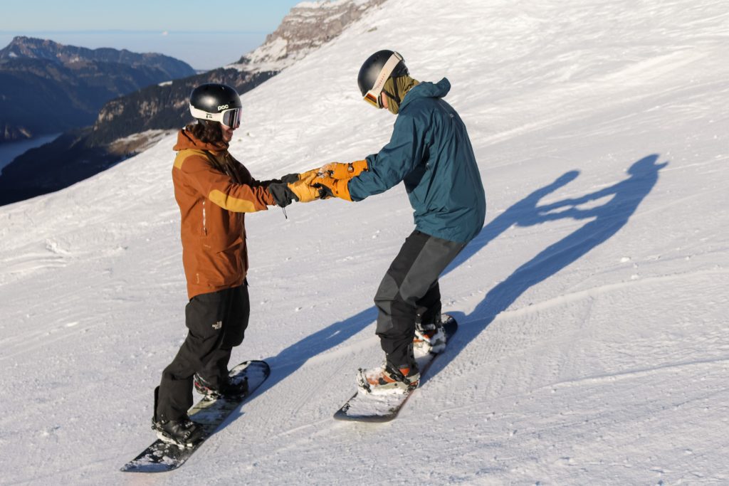Snowboard instructor supporting a student by holding their hands on a gentle slope in Engelberg