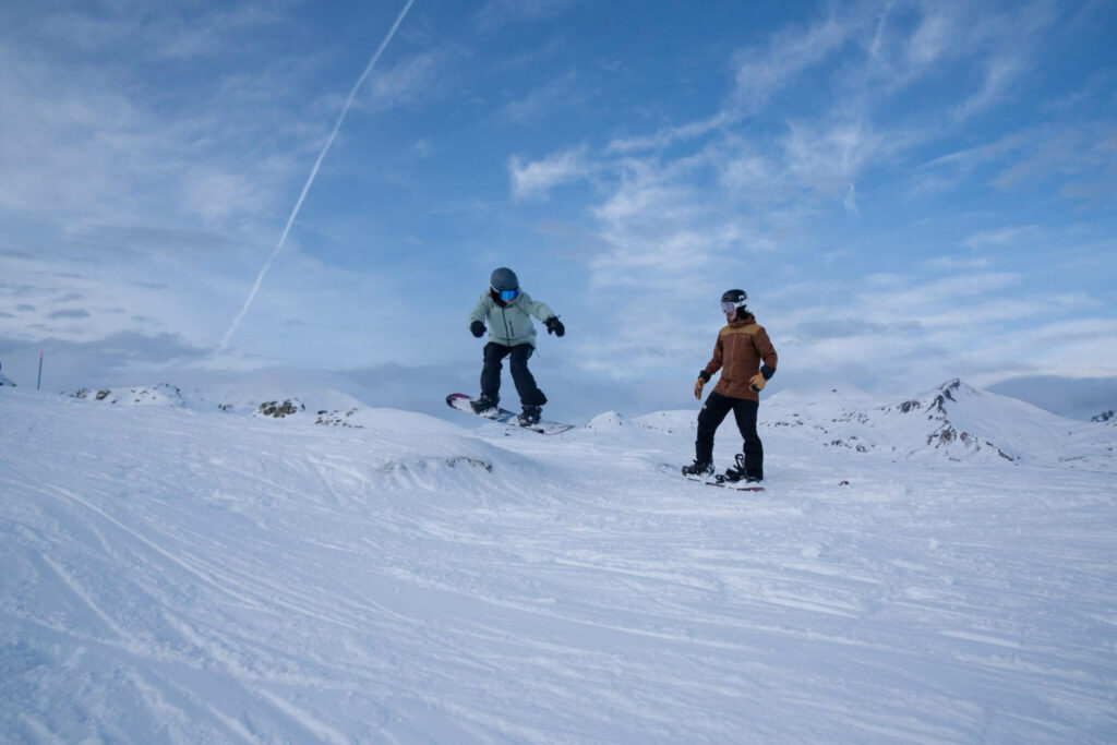 Snowboarder catching air off a small jump while the instructor watches, on the slopes above Engelberg
