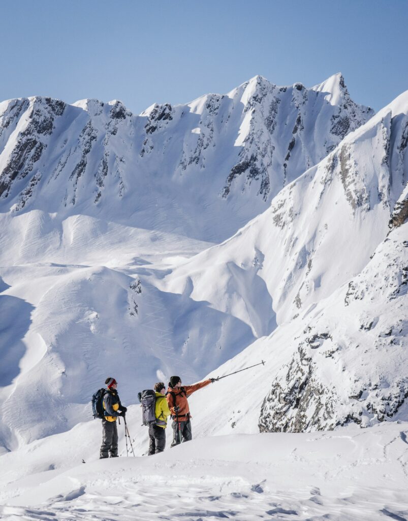 Three tourers with backpacks below a snowy mountain ridge, with one pointing towards the slope with a pole