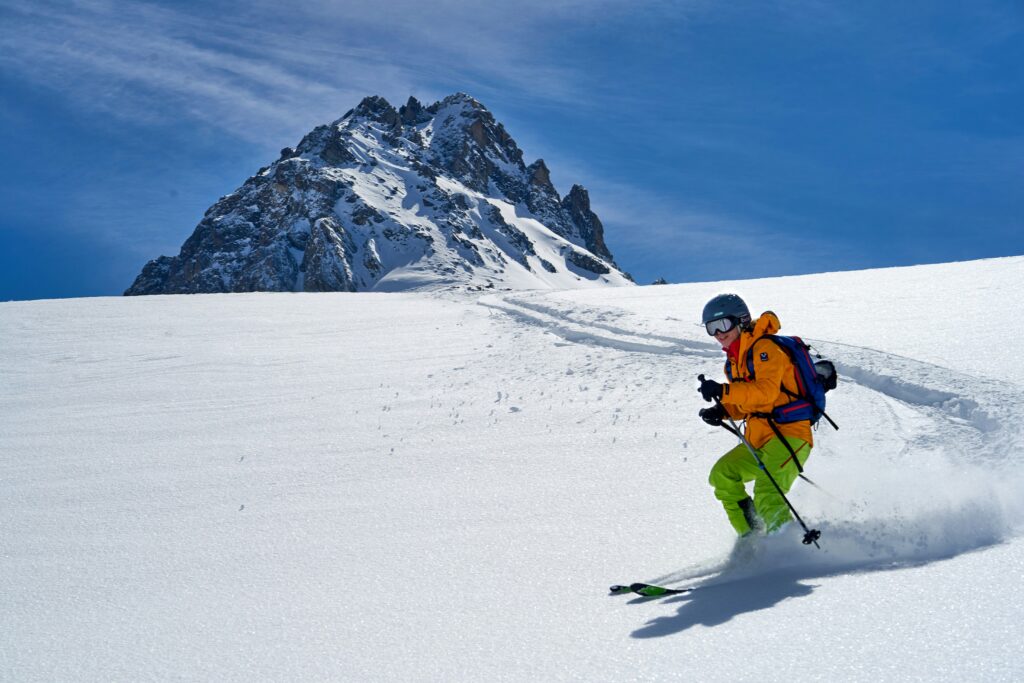 A skier in an orange jacket turning through fresh snow below a rocky peak
