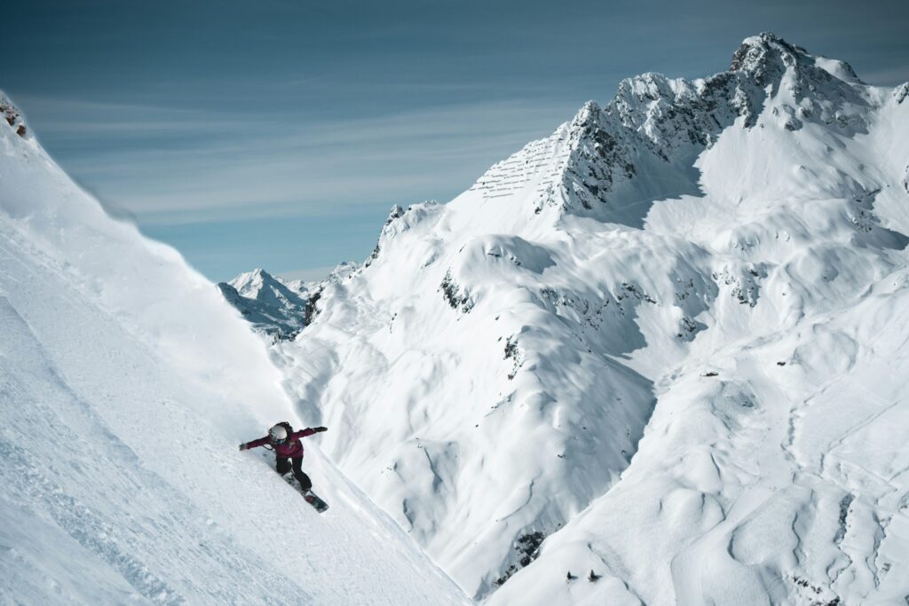 A snowboarder riding down a steep, untouched slope below snowy peaks